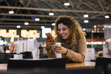 Zeitgenössische Dame in lässigen beigen Pullover mit Handy auf dem Tisch mit Kaffee zu gehen im Flughafen auf unscharfen Hintergrund - ADSF07479