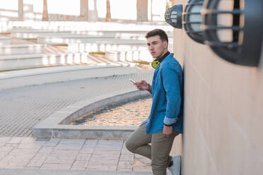 Side view of smart stylish man in bright headphones surfing mobile phone while leaning on marble wall in sunny day - ADSF07484