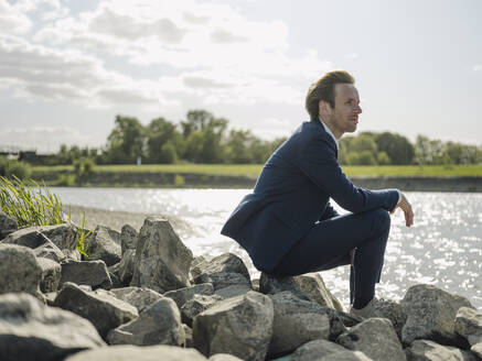 Nachdenklicher männlicher Unternehmer sitzt auf einem Felsen am Flussufer gegen den Himmel - GUSF04290