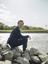 Thoughtful businessman sitting on rock at riverbank against sky during sunny day - GUSF04292