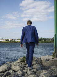 Businessman wearing suit walking on rocks at Rhine riverbank against sky - GUSF04296