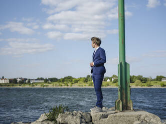 Geschäftsmann im Anzug steht auf einem Felsen am Flussufer gegen den Himmel - GUSF04297