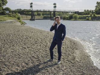 Businessman talking over smart phone while standing at riverbank during sunny day - GUSF04309