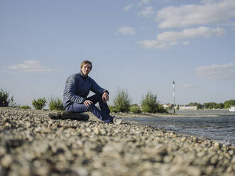 Mature man wearing warm clothing sitting at riverbank against sky on sunny day - GUSF04328