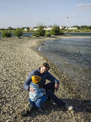 Father and daughter talking while sitting on log at riverbank against sky during sunny day - GUSF04330