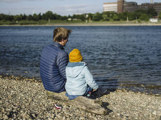 Vater und Tochter schauen auf den Rhein, während sie an einem sonnigen Tag auf einem Baumstamm sitzen - GUSF04331