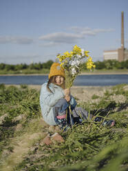 Lächelndes Mädchen hält Blumen und sitzt auf einer Wiese vor dem Himmel - GUSF04339