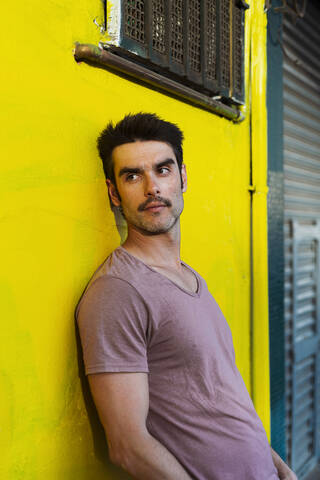 Thoughtful handsome man looking away while leaning on yellow wall in city stock photo