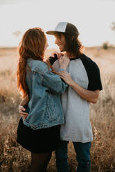 Young stylish teenagers embracing happily wile standing in remote rural field with warm sunset light - ADSF07539
