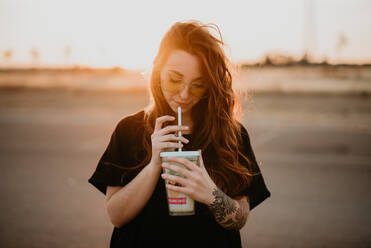 Charmante trendige Teenagerin mit Sonnenbrille und Tattoos, die einen Milchshake aus einem Glas mit Strohhalm bei Sonnenuntergang genießt - ADSF07543