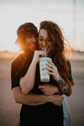 Young stylish teenagers embracing happily wile standing in remote rural field with warm sunset light drinking milkshake - ADSF07544