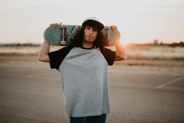 Stylish hipster teenage guy holding skateboard on behind head and looking at camera on empty road in sunset - ADSF07545