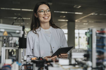 Smiling female scientist holding digital tablet looking away while standing by machinery in laboratory - MFF05905