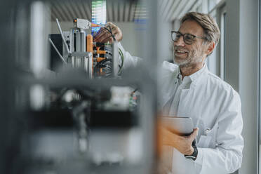 Smiling male scientist holding digital tablet inventing machinery in laboratory - MFF05921