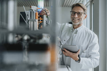 Smiling mature man with digital tablet inventing machinery in laboratory - MFF05922