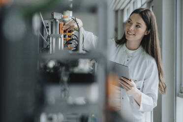 Smiling female scientist holding digital tablet inventing machinery in laboratory - MFF05923