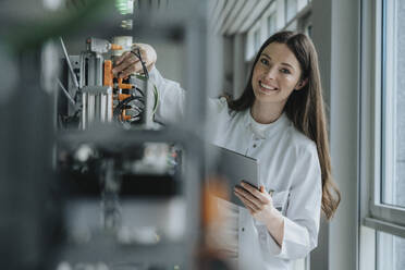 Smiling female scientist holding digital tablet inventing machinery in factory - MFF05924