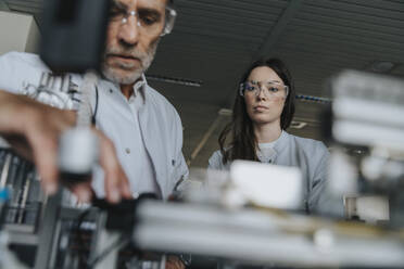 Männliche und weibliche Wissenschaftler mit Schutzbrillen bei der Untersuchung einer Maschine im Labor - MFF05946
