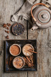 Draufsicht auf Masala Chai, serviert in Keramikschalen mit Sternanis und Zimtstangen, angeordnet auf einem Holztisch mit Teekanne und einem Stück Stoff - ADSF07650