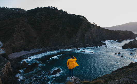 From above back view of person in vibrant yellow jacket standing on edge of cliff and enjoying amazing scenery of rocky sea coast during sunset in Spain - ADSF07657