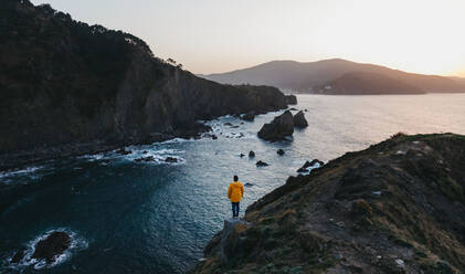 Von oben Rückansicht einer Person in leuchtend gelber Jacke, die am Rande einer Klippe steht und die herrliche Landschaft der felsigen Meeresküste bei Sonnenuntergang in Spanien genießt - ADSF07658