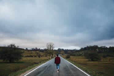 Rückenansicht eines anonymen Mannes in Freizeitkleidung, der auf einer leeren Asphaltstraße zwischen grünen Feldern mit bewölktem Himmel im Hintergrund geht - ADSF07661