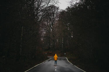 Rückenansicht eines anonymen Mannes in gelber Jacke, der auf einer leeren Asphaltstraße inmitten eines grünen Waldes geht - ADSF07665