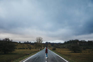 Side view of man in casual wear walking on empty asphalt road among green fields with cloudy sky on background - ADSF07666