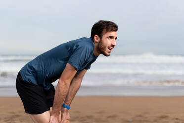 Seitenansicht eines bärtigen sportlichen Mannes in Sportkleidung, der nach einem langen Lauf am leeren Sandstrand eine Pause macht und wegschaut - ADSF07699