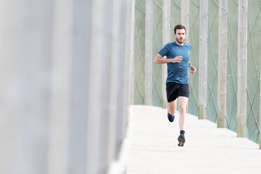 Highly motivated bearded male athlete in blue t shirt and shorts running outdoors under cover looking at camera - ADSF07702