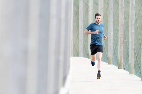 Hochmotivierter, bärtiger männlicher Sportler in blauem T-Shirt und Shorts, der im Freien läuft und in die Kamera schaut - ADSF07702