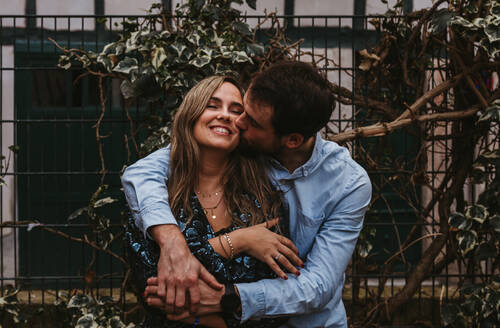 Zufriedener junger Mann und Frau, die sich umarmen und küssen, während sie in der Nähe eines Metallzauns auf einer Stadtstraße stehen und den Sommertag gemeinsam genießen - ADSF07708