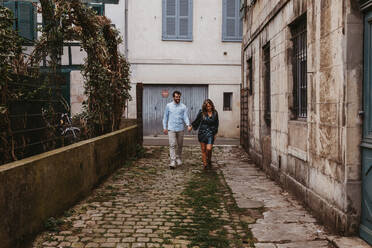 Fröhliches junges Paar in stilvoller Freizeitkleidung, das sich an den Händen hält und lächelt, während es auf einer alten engen Straße in der Stadt spazieren geht - ADSF07709