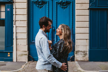 Seitenansicht eines glücklichen jungen Paares in legerer Kleidung, das sich umarmt und gegenseitig anschaut, während es vor einem alten Steingebäude mit blauen Türen auf einer Stadtstraße steht - ADSF07711