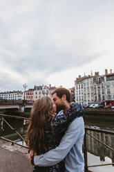 Seitenansicht eines glücklichen jungen zärtlichen Paares, das sich umarmt und küsst, während es auf einem Steindamm in der Nähe eines Flusses mit alten Gebäuden im Hintergrund in der Stadt Bayonne in Frankreich steht - ADSF07718