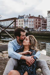 Positives junges Paar in Freizeitkleidung genießt romantisches Date, während sie zusammen auf Stein Grenze in der Stadt mit alten Gebäuden im Hintergrund sitzen - ADSF07719