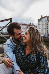 Positives junges Paar in Freizeitkleidung genießt romantisches Date, während sie zusammen auf Stein Grenze in der Stadt mit alten Gebäuden im Hintergrund sitzen - ADSF07720