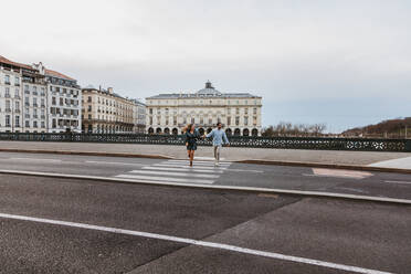 Glückliches junges romantisches Paar in stilvoller Kleidung lachend und Händchen haltend beim Überqueren einer Brücke mit historischen Gebäuden im Hintergrund während einer Stadtbesichtigung in Bayonne in Frankreich - ADSF07721