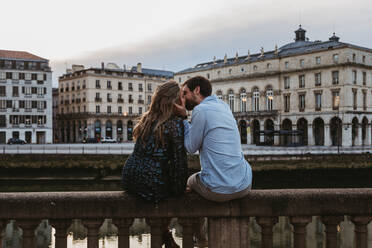 Back view of happy couple sitting kissing each other on old stone fence spending summer evening together in Bayonne - ADSF07723