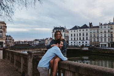 Happy young female sitting on old stone fence and embracing affectionate boyfriend while spending summer evening together in Bayonne - ADSF07724