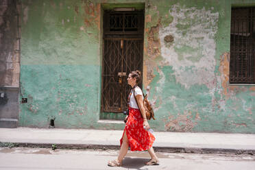 Seitenansicht der Frau im Urlaub in bunten roten Rock mit Rucksack und Fotokamera zu Fuß entlang der Straße mit alten Gebäude auf dem Hintergrund in Kuba - ADSF07726
