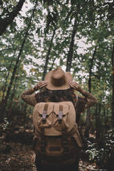 Back view of anonymous woman with backpack adjusting hat while traveling through green forest in nature - ADSF07728