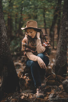 Calm young female in stylish hat and scarf sitting on ground with closed eyes and resting in green forest - ADSF07729