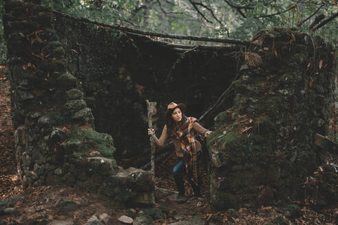 Woman with wooden stick exploring old ruins in green forest in nature - ADSF07733