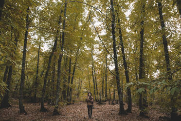 Young woman in hat wrapping in checkered scarf while standing on dry leaves in autumn forest - ADSF07736