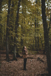 Seitenansicht einer jungen Frau mit Hut, die sich in einen karierten Schal einwickelt, während sie auf trockenen Blättern im Herbstwald steht und nach oben schaut - ADSF07737