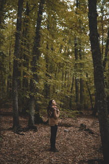 Side view of young woman in hat wrapping in checkered scarf while standing on dry leaves in autumn forest looking up - ADSF07737