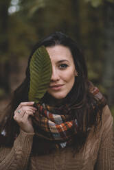 Stilvolle Dame in Strickpulli und kariertem Schal, die ihre Augen mit einem Blatt bedeckt, während sie vor einem unscharfen Waldhintergrund steht - ADSF07738