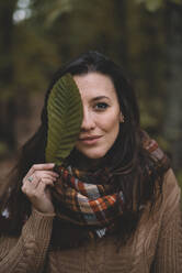 Stylish lady in knitted sweater and checkered scarf covering eye with leaf while standing on blurred background of forest - ADSF07738