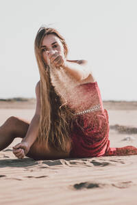 Attraktive junge Frau mit langen blonden Haaren in einem schicken roten Kleid sitzt an der Küste und gießt Sand aus der Hand, während sie in die Kamera schaut - ADSF07742